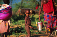 Naked Samburu child follows his mother home. She holds a container of maize she has received from aid workers with EdFri International