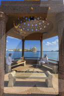 Seated Omani men wearing traditional dishdasha on Mutrah Corniche, with view to Al Said Royal Yacht, Muscat, Oman, Middle East