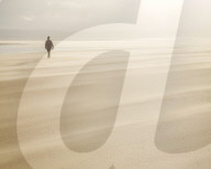 Man walking across a windy beach with dry shifting sands creating a cloud underfoot