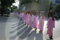 Long line of Buddhist nuns dressed in pink robes, heads covered, alms bowls on their heads, crossing main road in Mandalay.