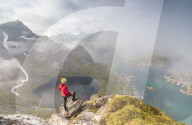 Hiker on summit admires the blue lake and sea framing the village, Reinebringen, Moskenesoya, Lofoten Islands, Norway, Scandinavia, Europe