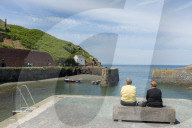 Two ladies sat on a bench at Porthgain harbour as the Pembrokeshire coast path extends up the cliffs beyond.