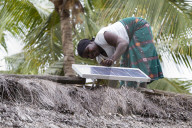A woman, known as a Solar Mama, fixes a solar panel to the top of a traditional thatched mud hut.