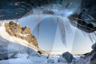View from ice cave towards sunlit mountains with icicles hanging from cave entrance, near Jokulsarlon, South Iceland