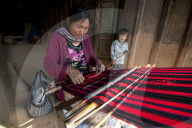 Woman weaving traditional red Naga shawl, young daughter beside her, on the verandah of the family home, Nagaland