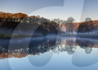 Dawn light reveals the autumn colour of trees and distant fells with mist lying above Elterwater revealing a perfect reflection