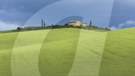 A single Tuscan farmhouse sits perched on top of a hill beyond verdant rolling green fields of the Val d'Orcia