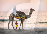 Boy stares in amazement at Arabic man wearing a thawb riding a camel on a beach, Dubai, UAE