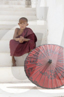 Young monk dressed in red, sits on steps with red parasol at the Myatheindan Pagoda (White Temple) in Mingun, Myanmar (Burma)