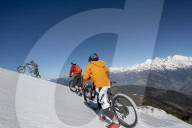 Mountain bikers cycle along a snow covered slope in the Himalayas with views of the Langtang range in the distance