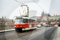 A traditional red tram crosses MÃ¡nesÅ¯v most (bridge) with St. Vitus Cathedral and Prague Castle in the background.