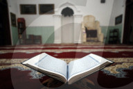 An open Holy Quran on wood stand with mihrab in background.