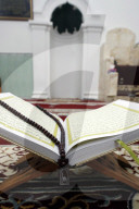 An open Holy Quran and a muslim prayer beads on wood stand with mihrab in background.