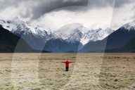 Man in red coat stood holding out his arms looking at snow capped mountains, Fiordland National Park, New Zealand