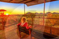 Tourist woman sitting on a bench at platform dune viewing area and looking Mount Olga in Uluru-Kata Tjuta National Park at orange sunset sky. Australian outback in Northern Territory, Australia