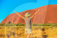 Carefree tourist woman with raised arms enjoys Uluru Ayers Rock at sunset in Uluru-Kata Tjuta National Park. Blonde traveler in Australian outback Red Centre. Tourism in Northern Territory, Australia.