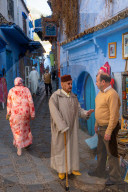 Street scene: old man with stick and red fez (hat) stops to talk with a the owner of a bookshop, Chefchaouen, Morocco
