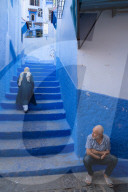 Woman in dark blue traditional clothes climbing up the stairs of alleyway, man seated in the foreground, in Chefchaouen, Morocco