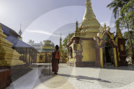 A monk in maroon robes holding a staff and a steel alms bowl standing in the grounds of a temple