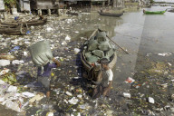 Two men unloading sacks of produce from a small boat in Sittwe harbour, wading through polluted water full of plastic rubbish