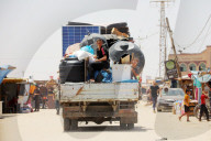 Palestinians  ride in the back of a truck loaded with solar panels, cisterns, and other items moving past the tents and shelters